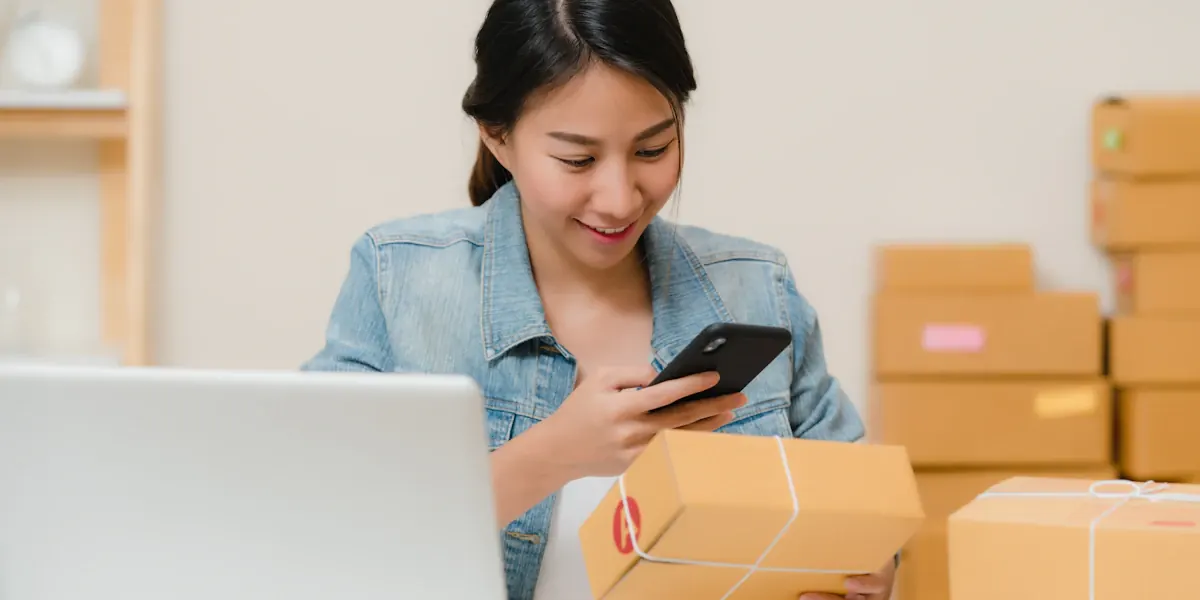 Online seller checking a parcel with her phone next to a laptop and boxes