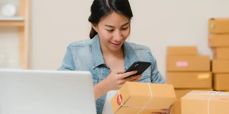 Online seller checking a parcel with her phone next to a laptop and boxes